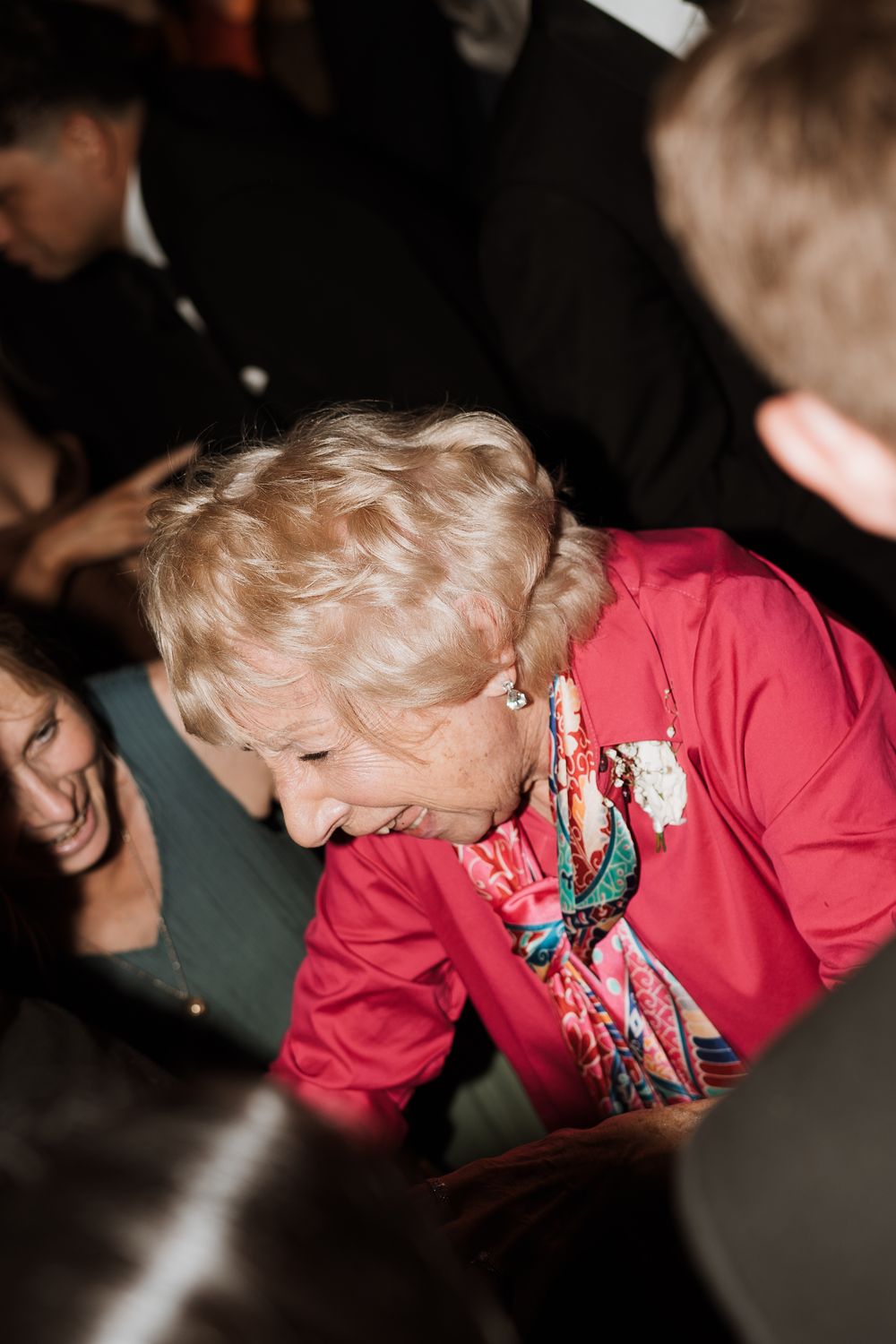 Someone in a bright red sweater with a colorful scarf leans in while laughing during a dimly lit gathering.