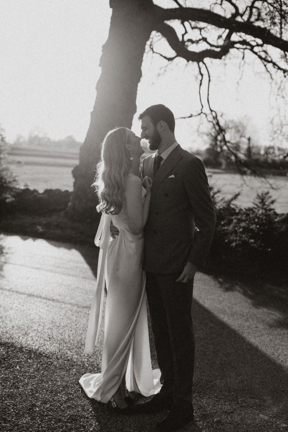 A romantic black and white portrait of a couple embracing under a tree at sunset.
