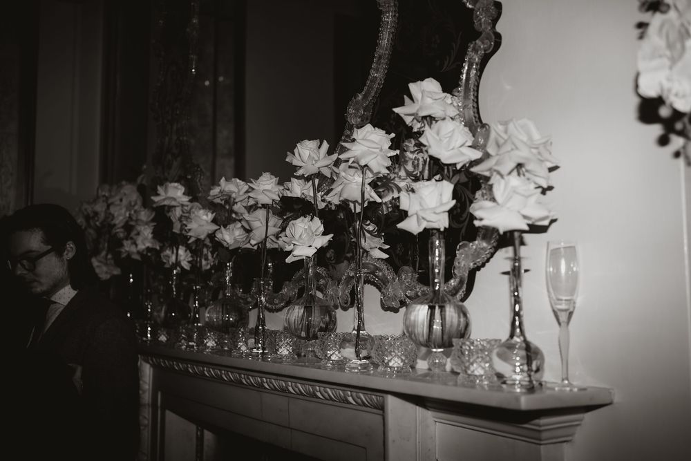 A black and white photo of elegant floral arrangements with white roses and champagne glasses on a mantelpiece.
