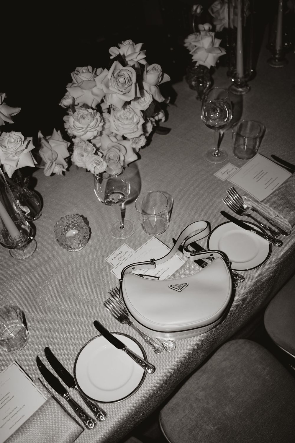 Black and white photo of elegant table setting with rose centerpiece, wine glasses, plates and silverware at formal event.