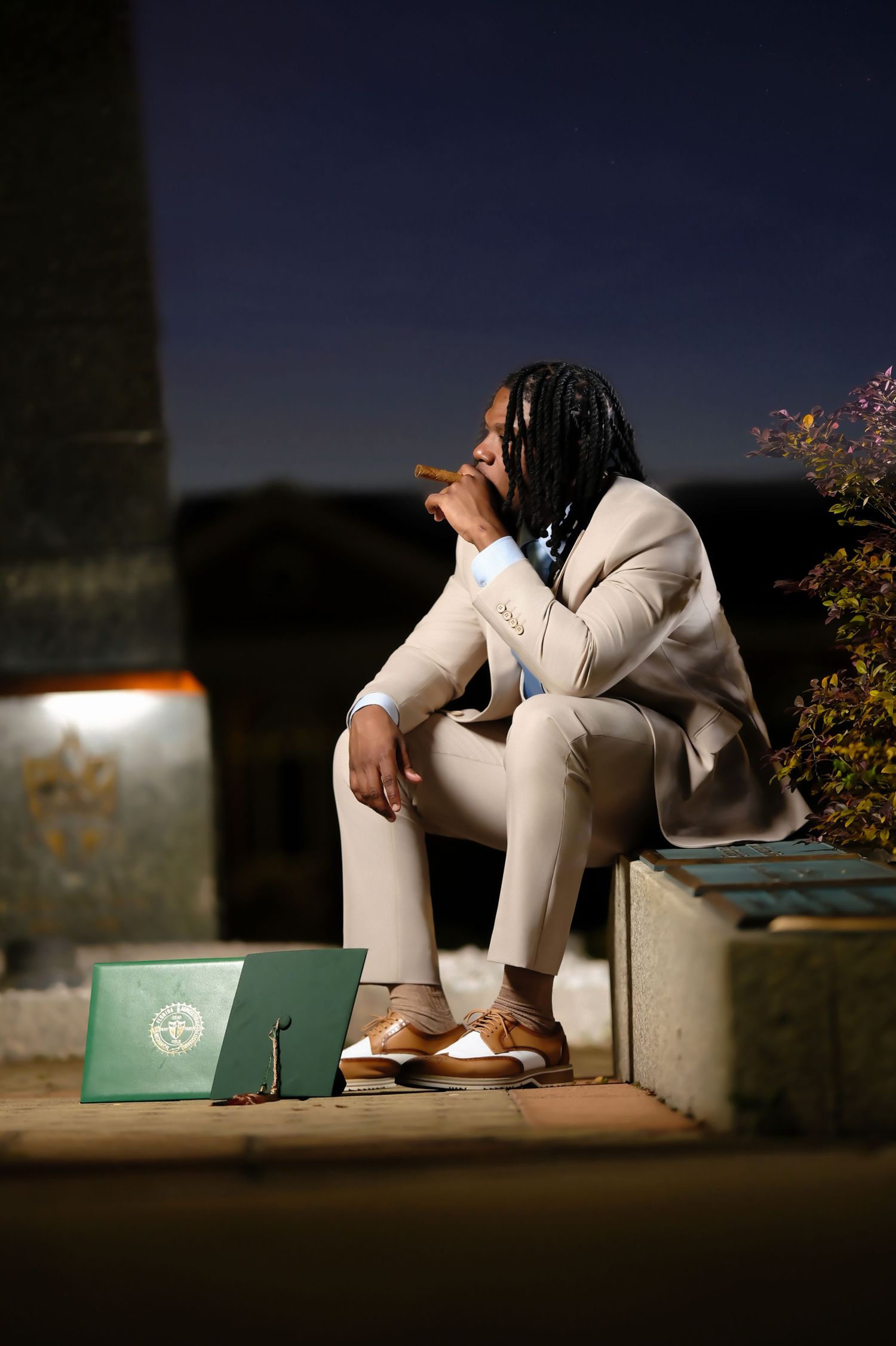Fashion portrait of someone in a stylish white suit sitting thoughtfully outdoors at night with a shopping bag nearby.