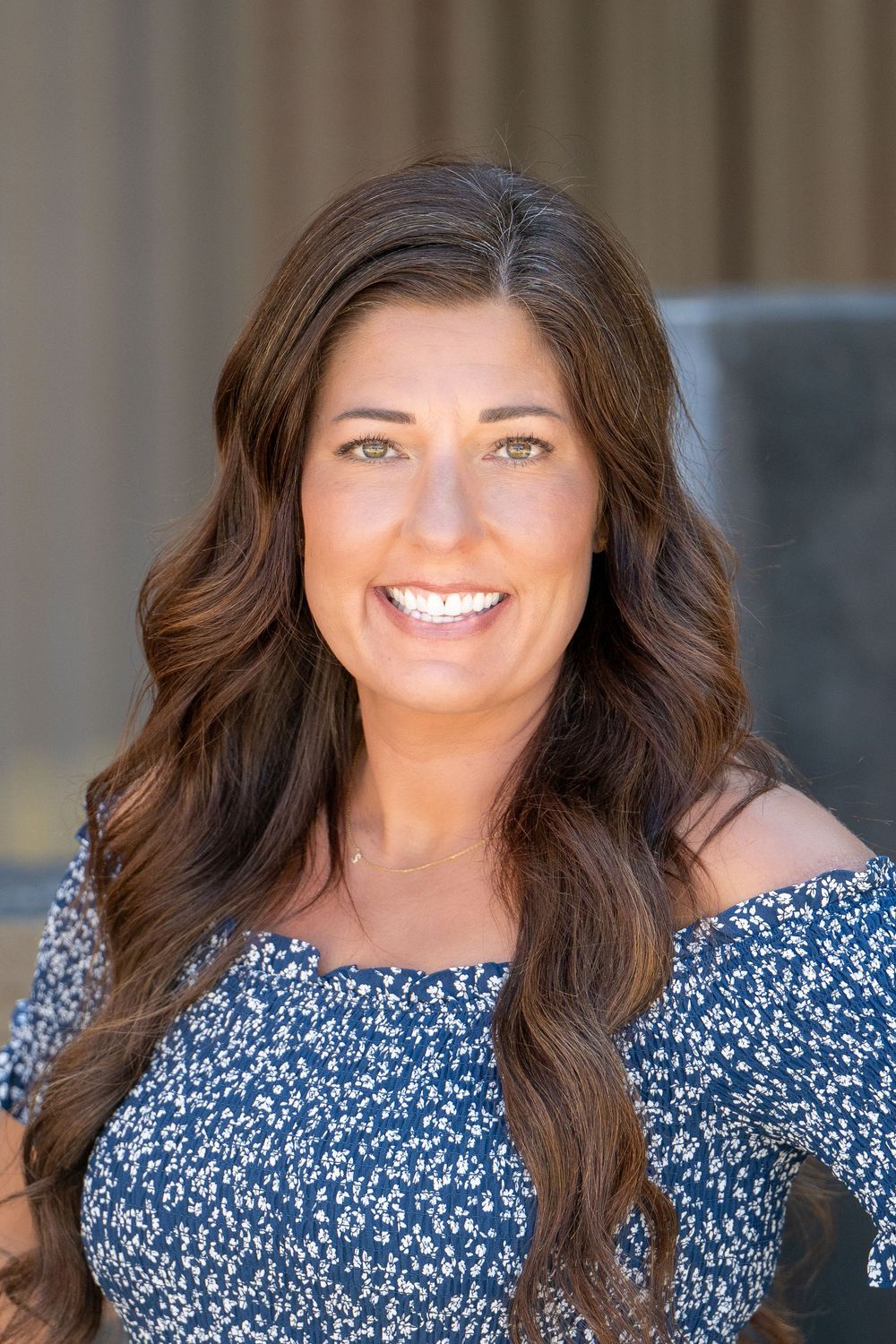 Professional headshot portrait of someone with long wavy brown hair wearing a blue floral off-shoulder top against neutral background.