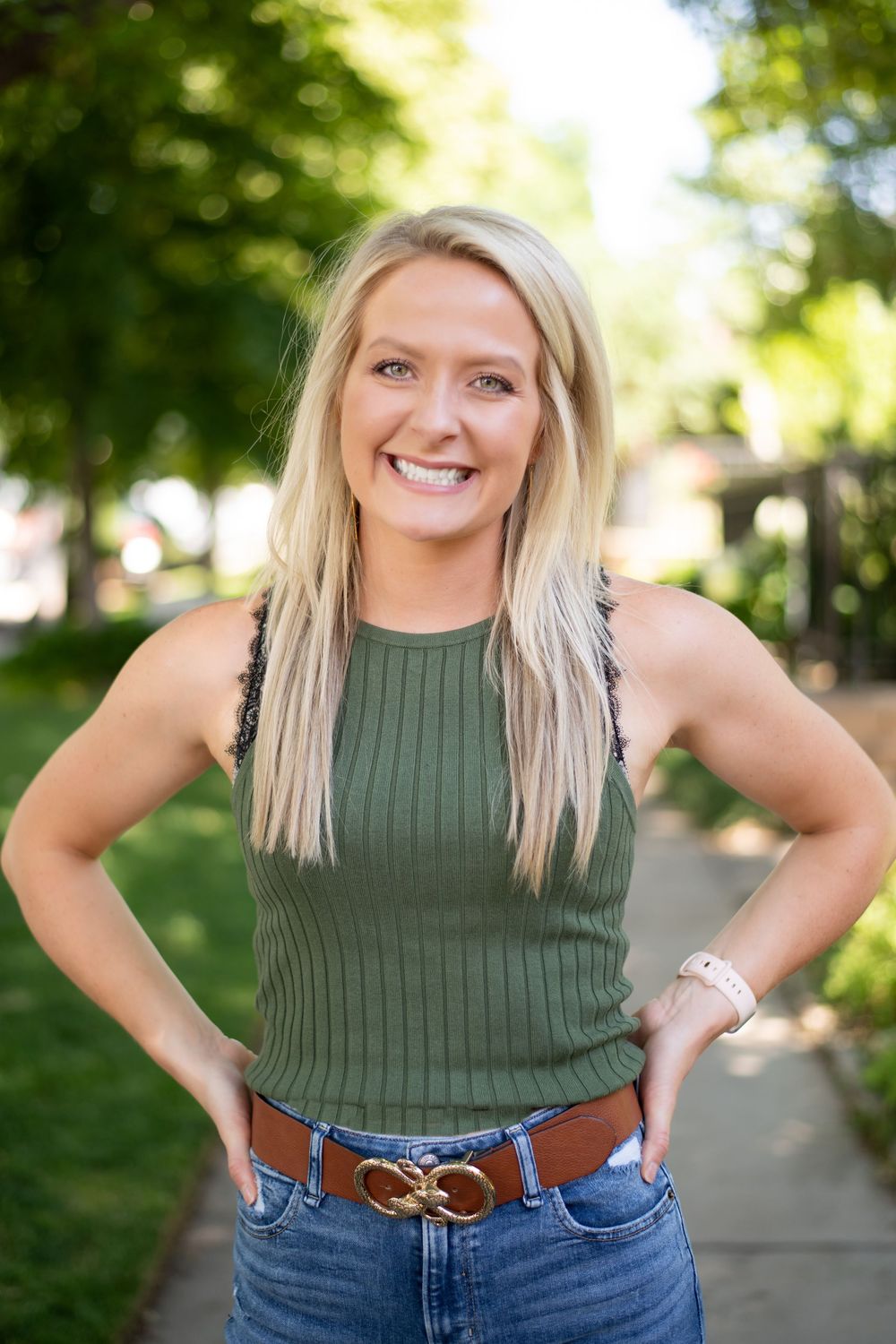 Portrait of someone in a green ribbed tank top and jeans with a brown belt standing outdoors in a sunny park setting.