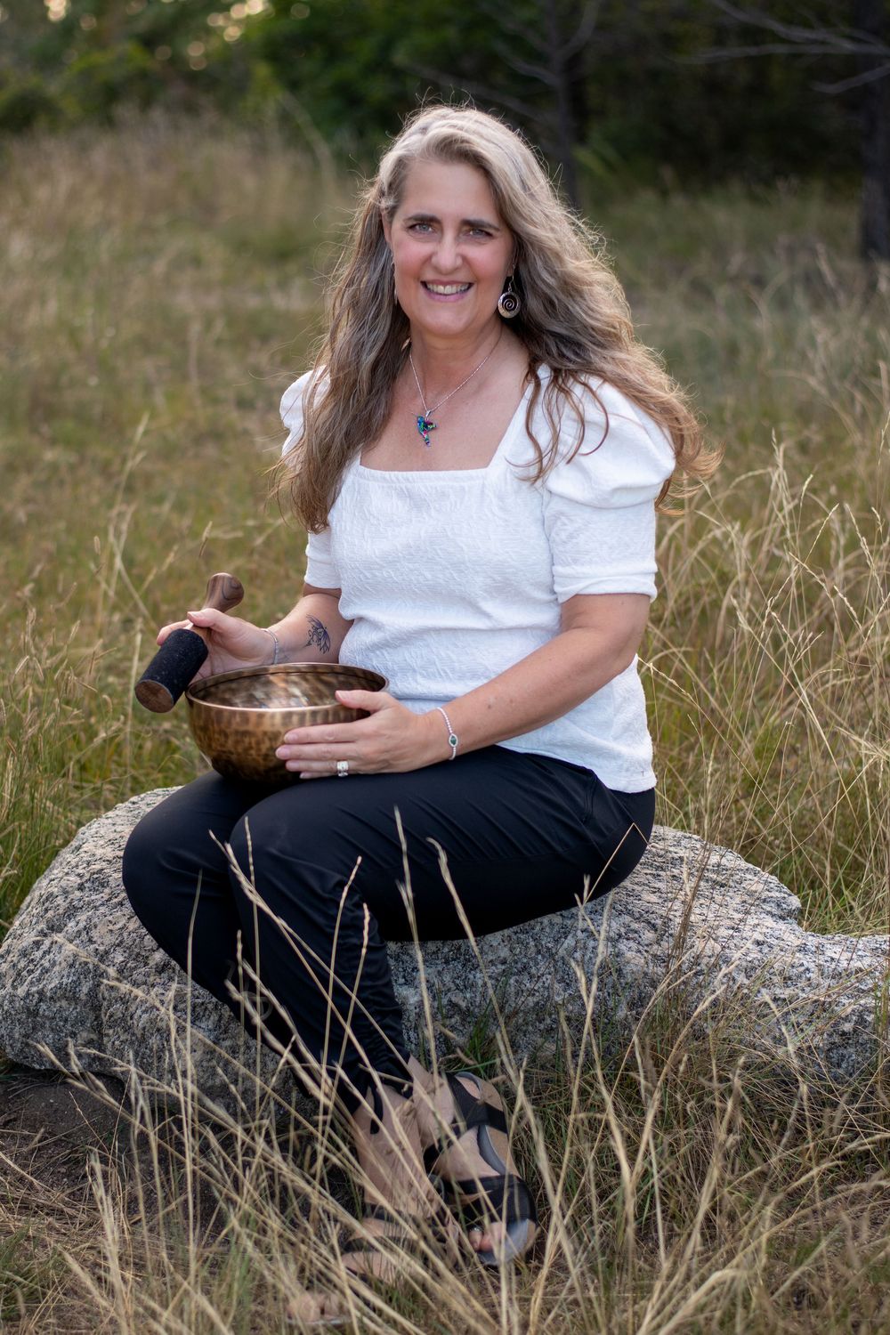 Person wearing white top sits on rock in grassy field while holding a meditation singing bowl.