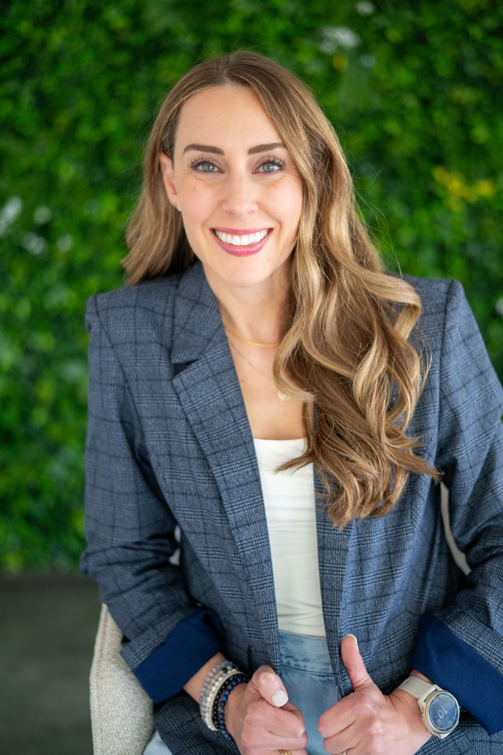 Professional headshot of someone in a navy blazer and white top against a green leafy background.