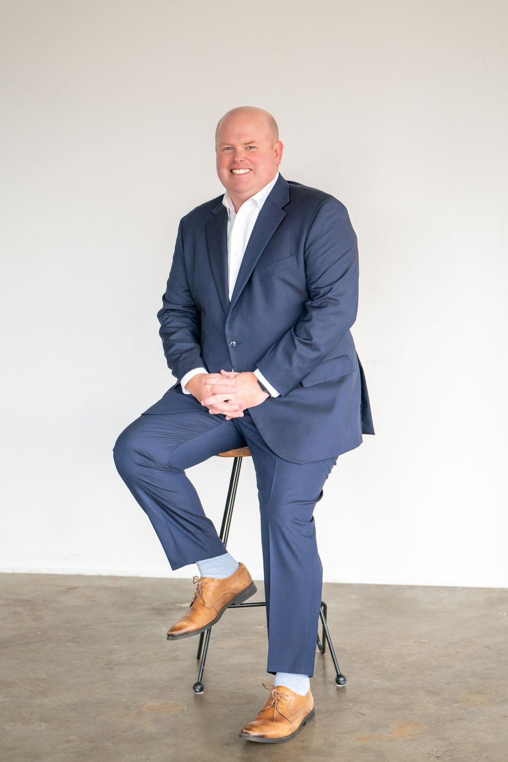 Professional business portrait of person in navy suit seated on stool against light background.