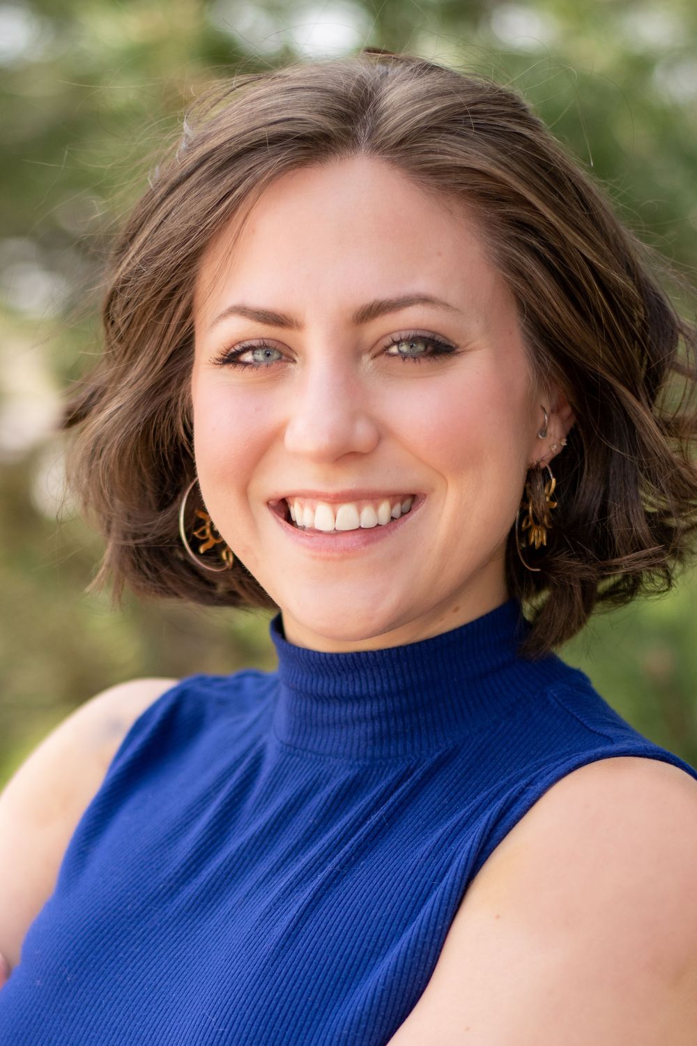 A person in a navy blue sleeveless turtleneck smiles warmly at the camera with gold hoop earrings and natural makeup.
