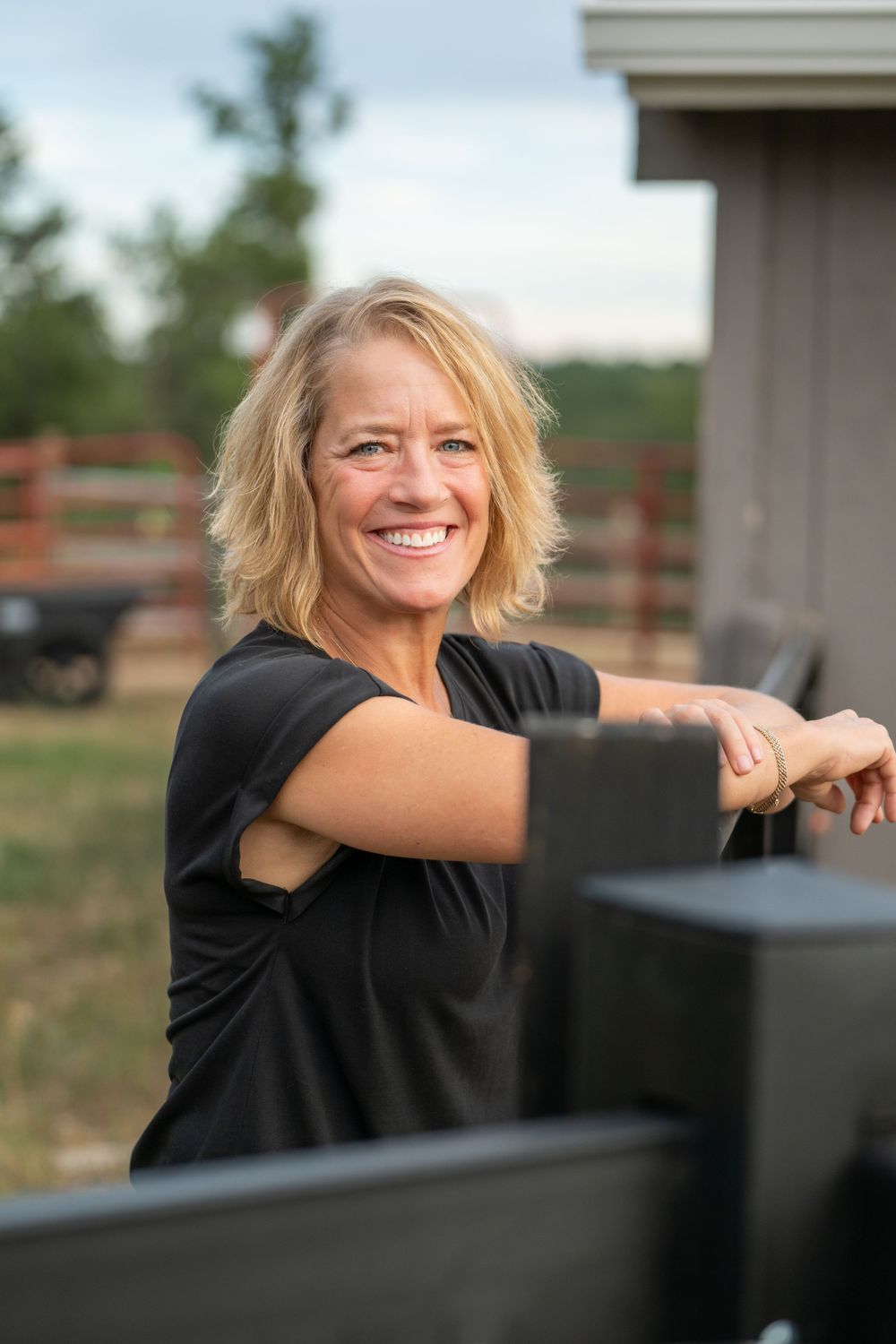 Smiling woman in black shirt leaning on outdoor railing with natural background on sunny day.