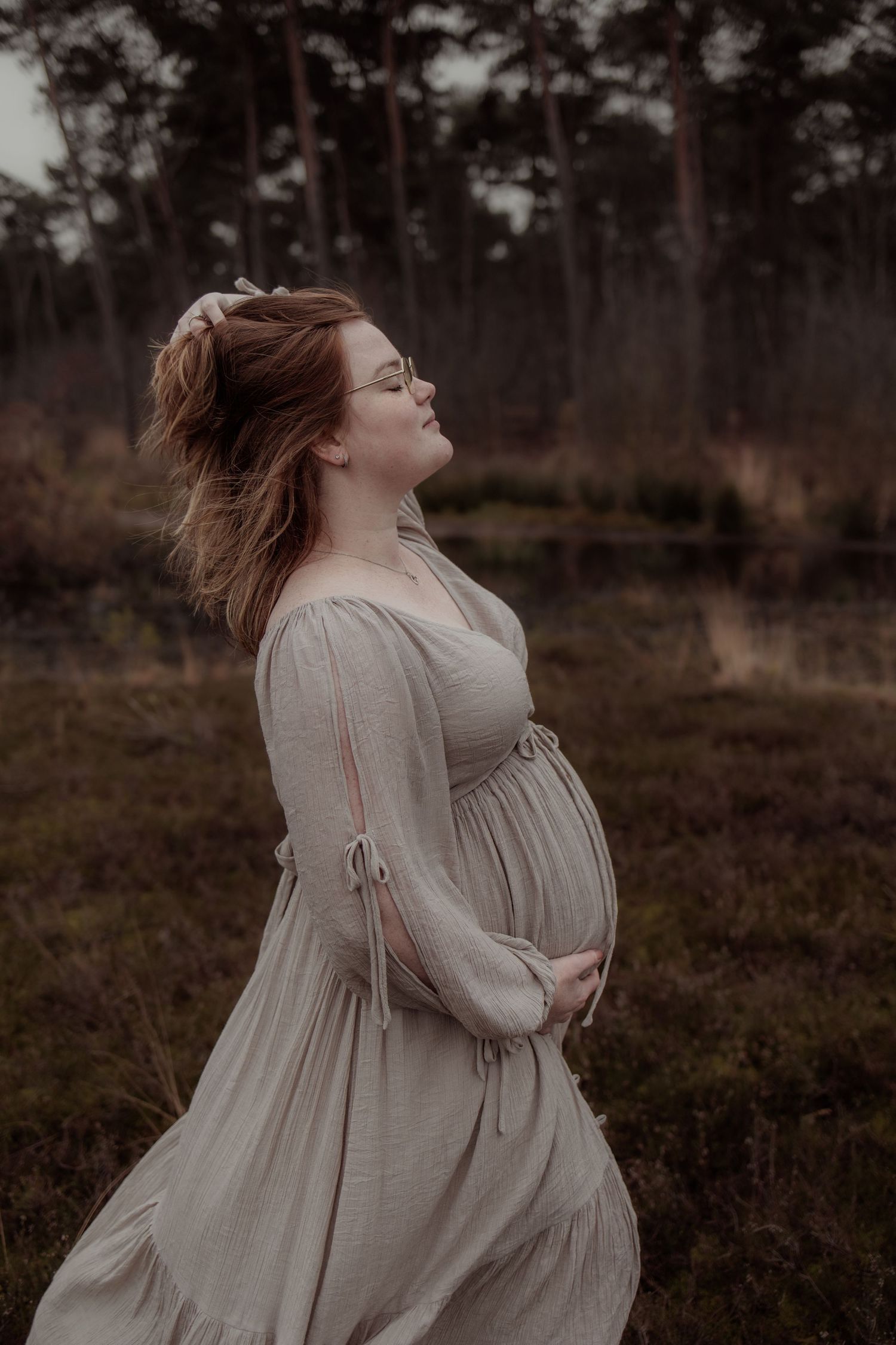 Ethereal maternity photo of a person in a flowing white dress standing in a moody forest setting at dusk.