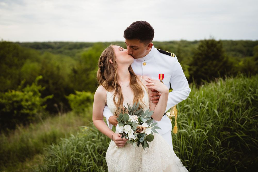 A couple shares a romantic moment among tall grass with a bouquet of white flowers and greenery.