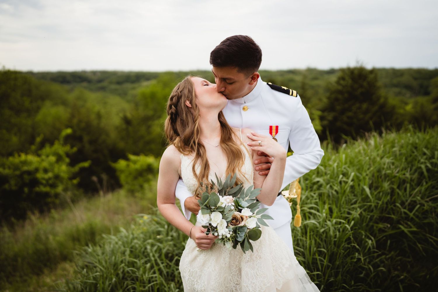 A couple shares a romantic moment among tall grass with a bouquet of white flowers and greenery.