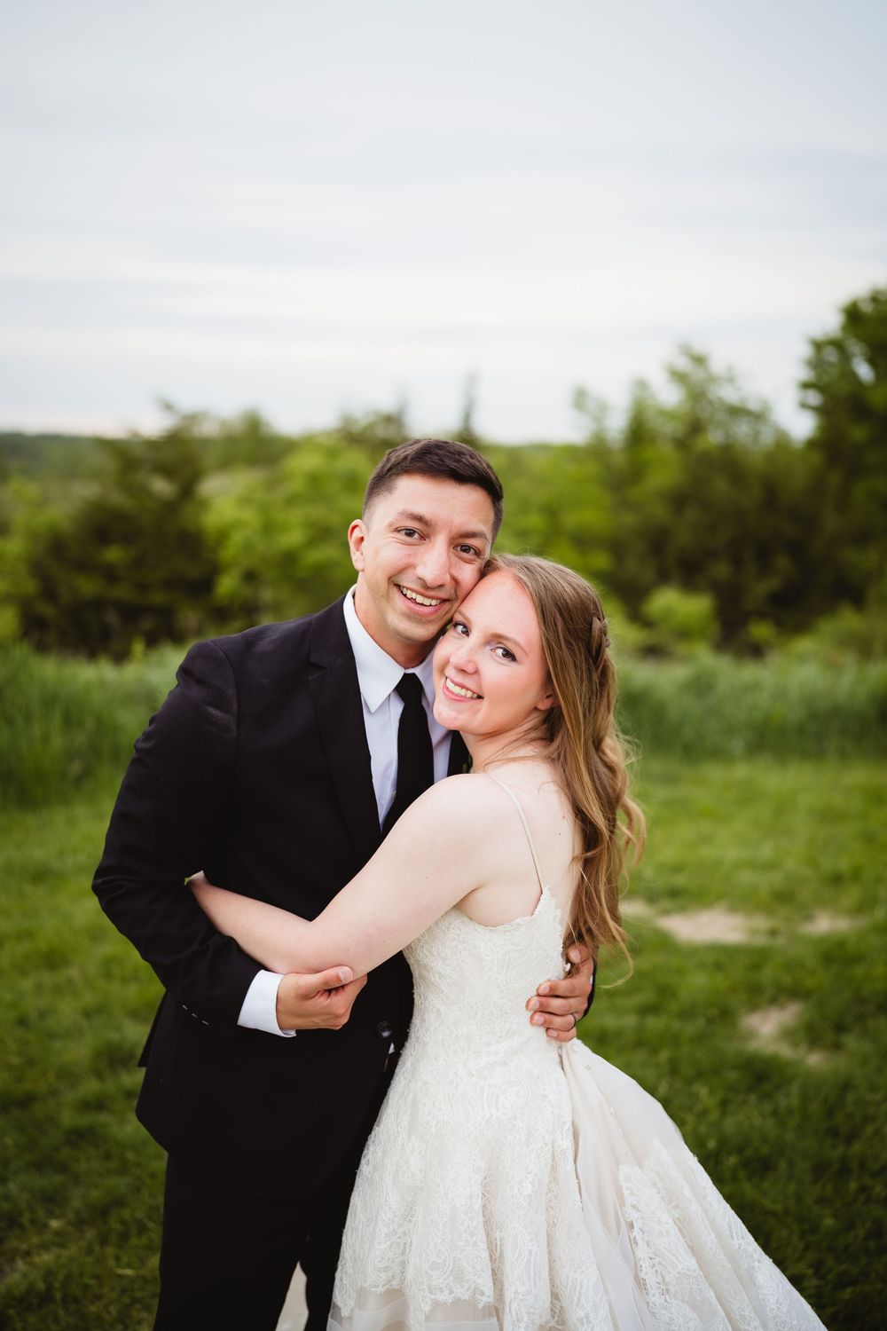A bride and groom embrace outdoors in a natural setting with green foliage and grass in the background.