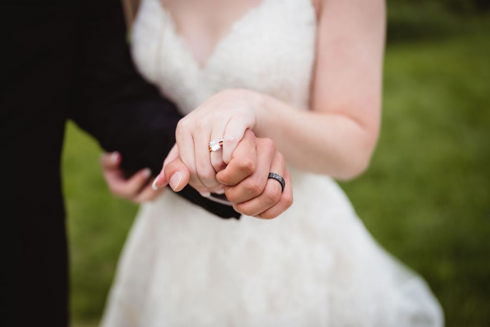 Close-up of newlyweds holding hands outdoors showing wedding rings and white bridal gown.