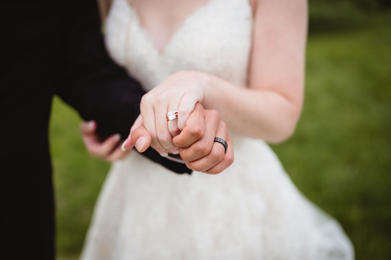 Close-up of newlyweds holding hands outdoors showing wedding rings and white bridal gown.