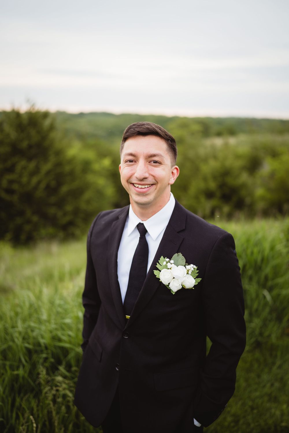 A groom in a black suit with white boutonniere smiles while standing in a lush green outdoor setting.