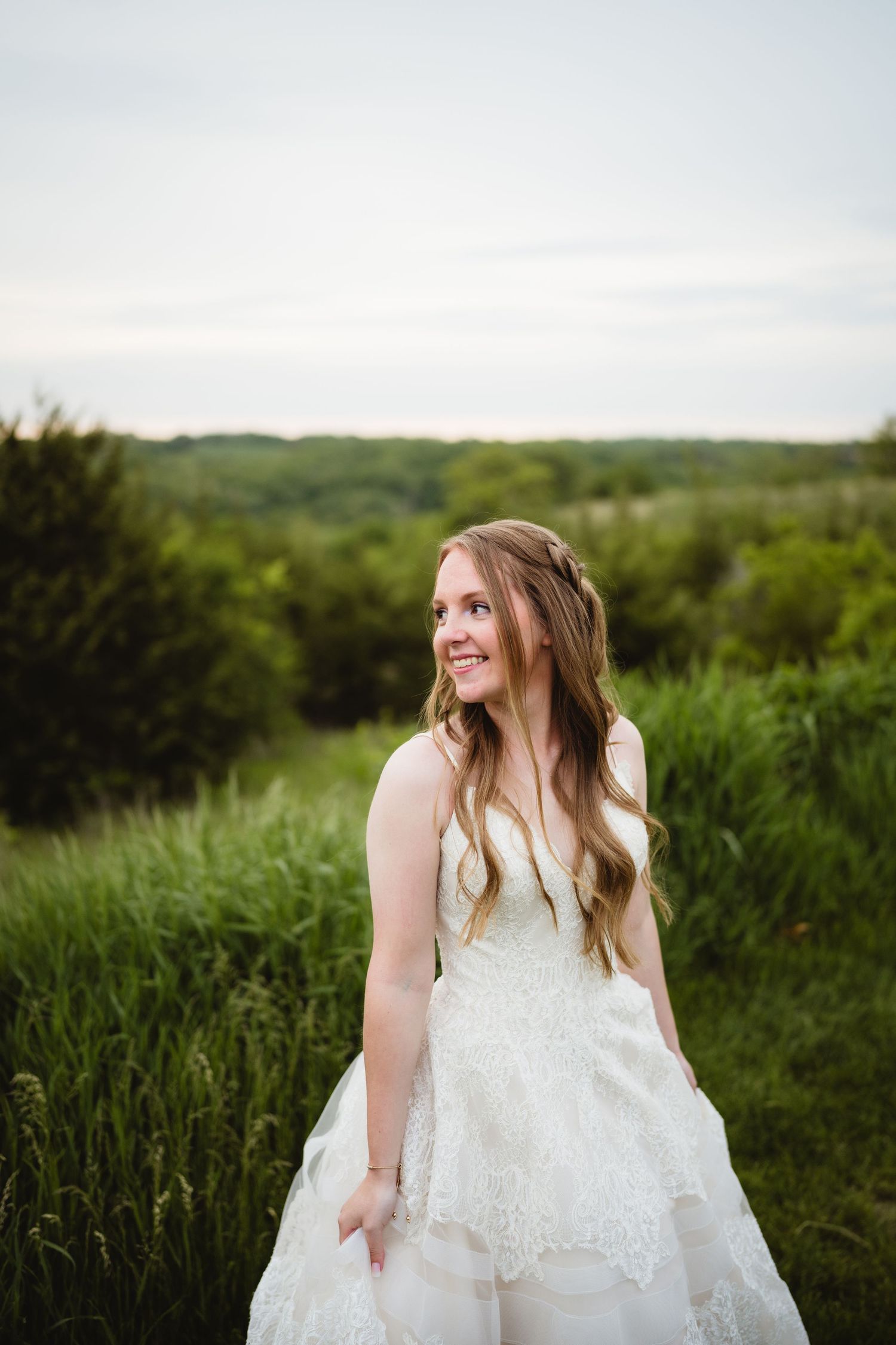 A bride in a white lace wedding gown poses in a field of tall grass with rolling hills in the background.