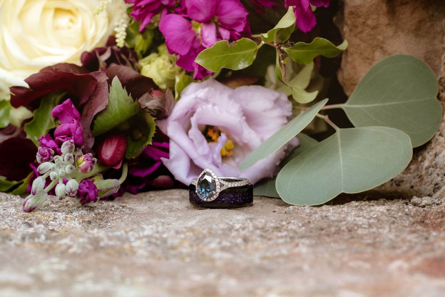 Diamond engagement ring resting on a rock surrounded by colorful purple and cream wedding bouquet flowers.