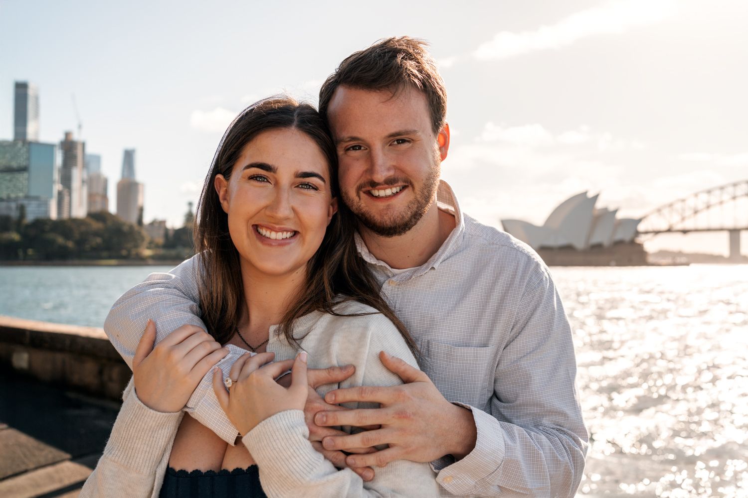 man hugs his girlfriend and both of them looking into the camera during a secret proposal photoshoot in Sydney golden hour