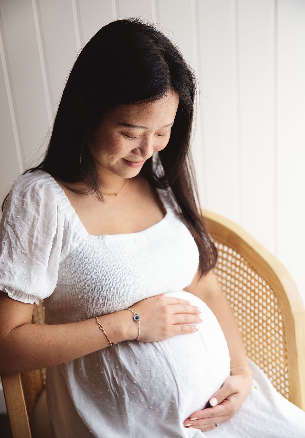A pregnant person in a white dress smiles while tenderly holding their baby bump in front of a cane chair. During a photoshoot in London