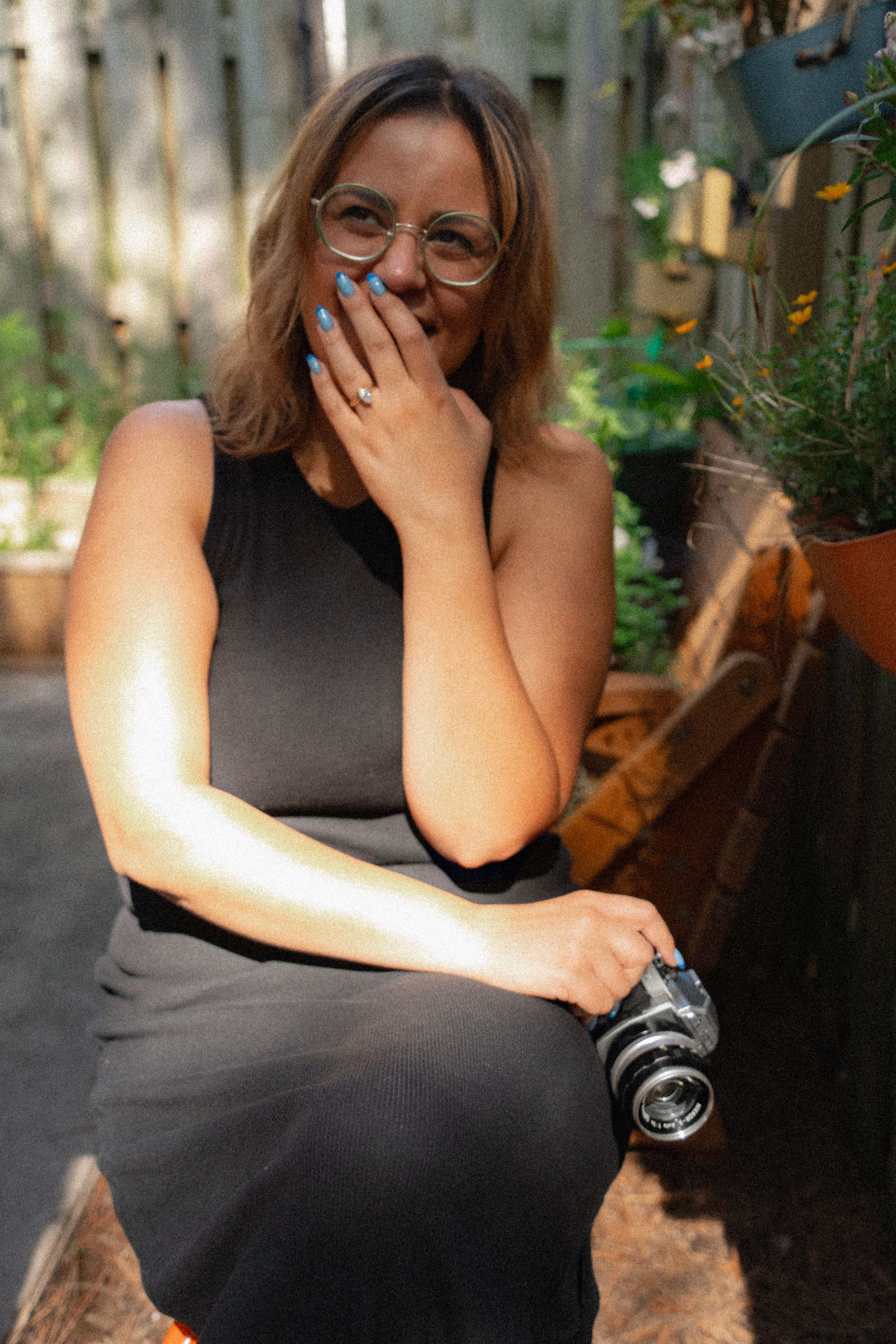A photographer in glasses wearing a black dress sits outdoors with a vintage camera while laughing.