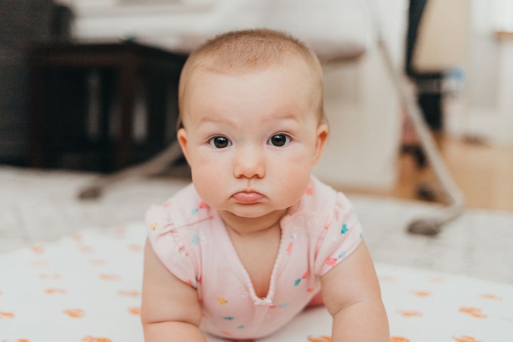 Baby wearing a pink onesie lies on patterned bedding at home during tummy time.