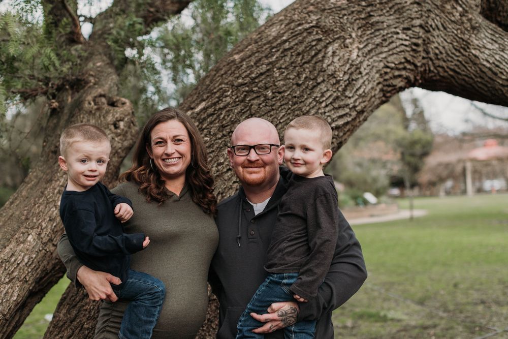 Happy family of four poses together by a large tree trunk in an outdoor setting.