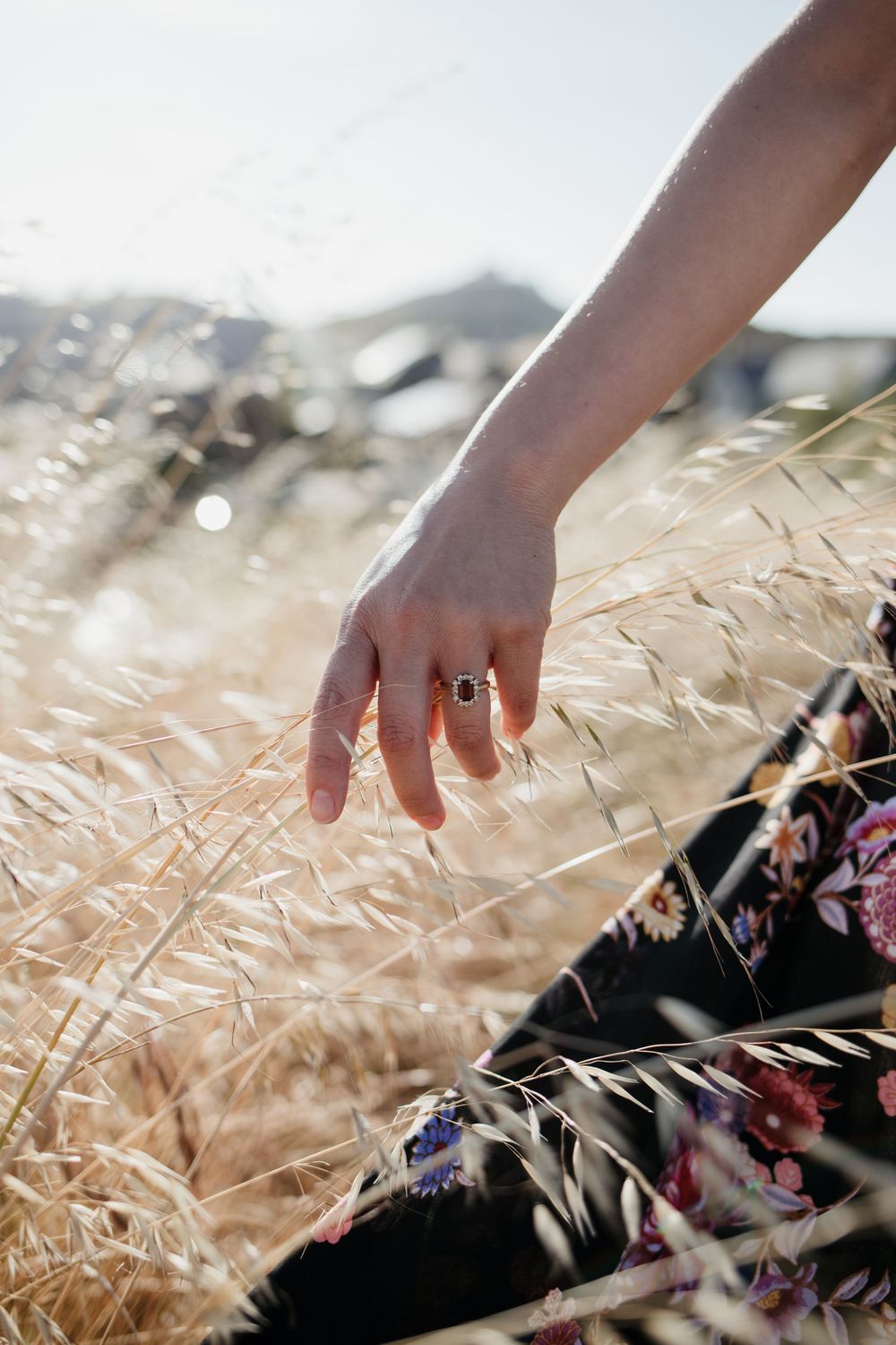 Hand wearing silver ring gently touches golden wheat stalks in sunlit field with blurred floral pattern in foreground.