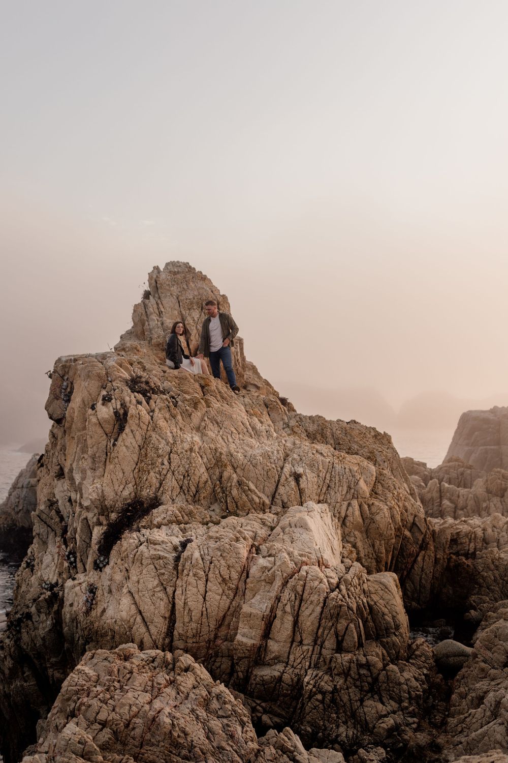 Two people sit atop a dramatic rocky cliff formation against a misty sunset sky along a rugged coastline.