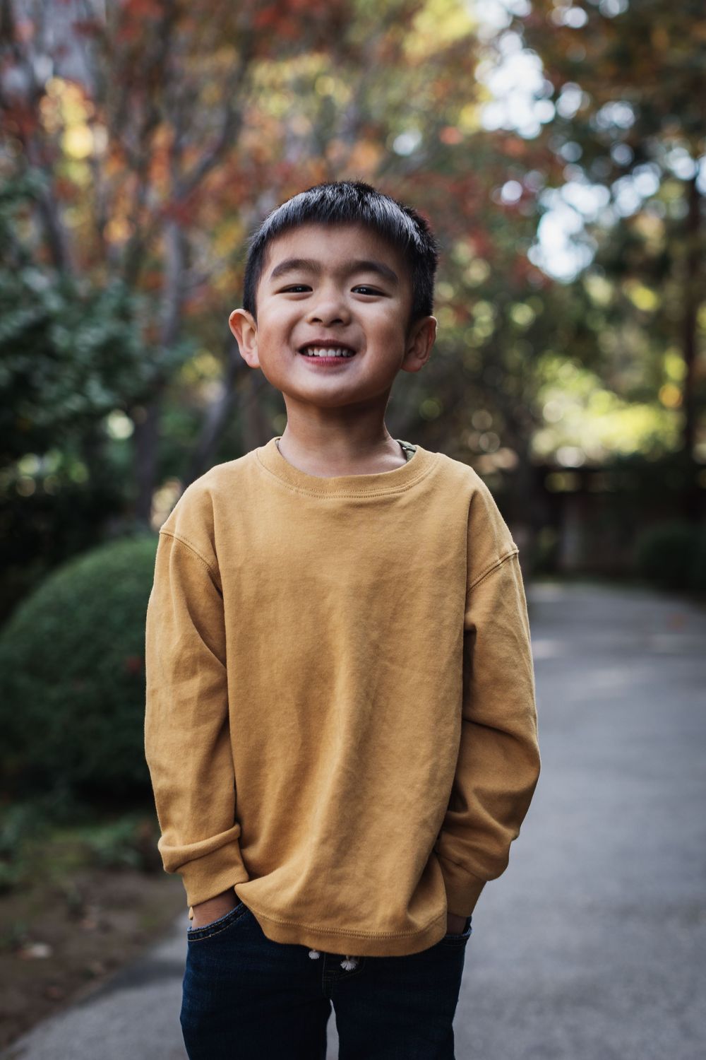 A child wearing a mustard yellow sweatshirt smiles joyfully while standing on a path surrounded by autumn foliage.