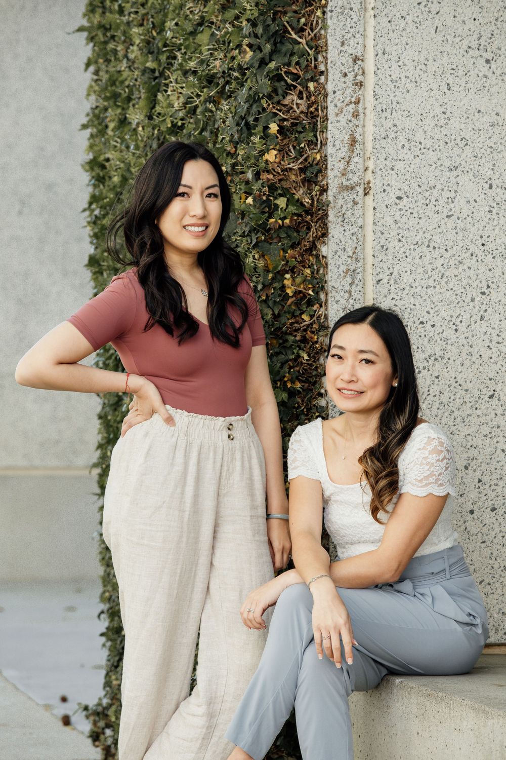 Two people wearing casual outfits pose together against a wall with climbing vines outdoors during daytime.