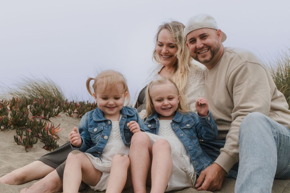 A family in casual clothing sits together outdoors on sandy ground with tall grass in the background.