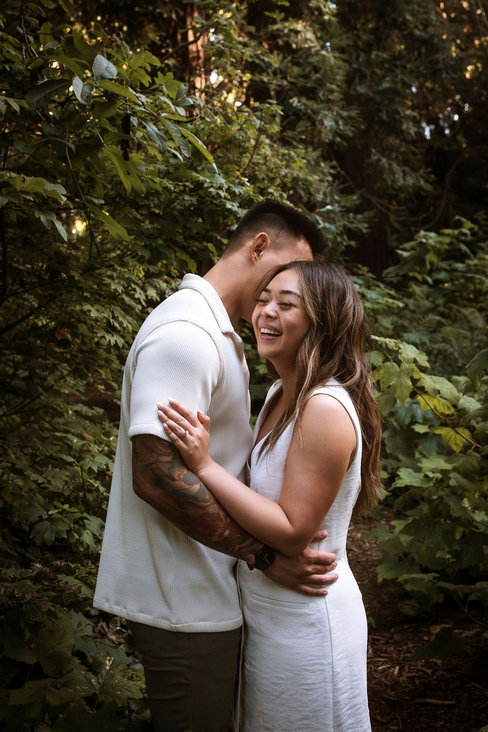 A romantic couple in white outfits share an intimate embrace while laughing together in a lush green forest setting.