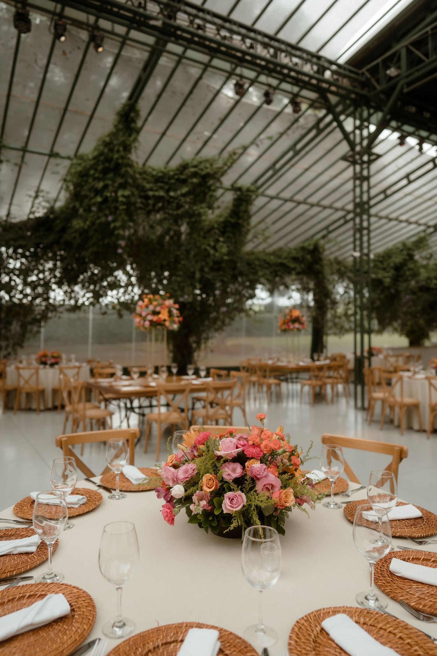 Wedding reception venue with greenhouse ceiling, wooden chairs and floral centerpieces on round tables.