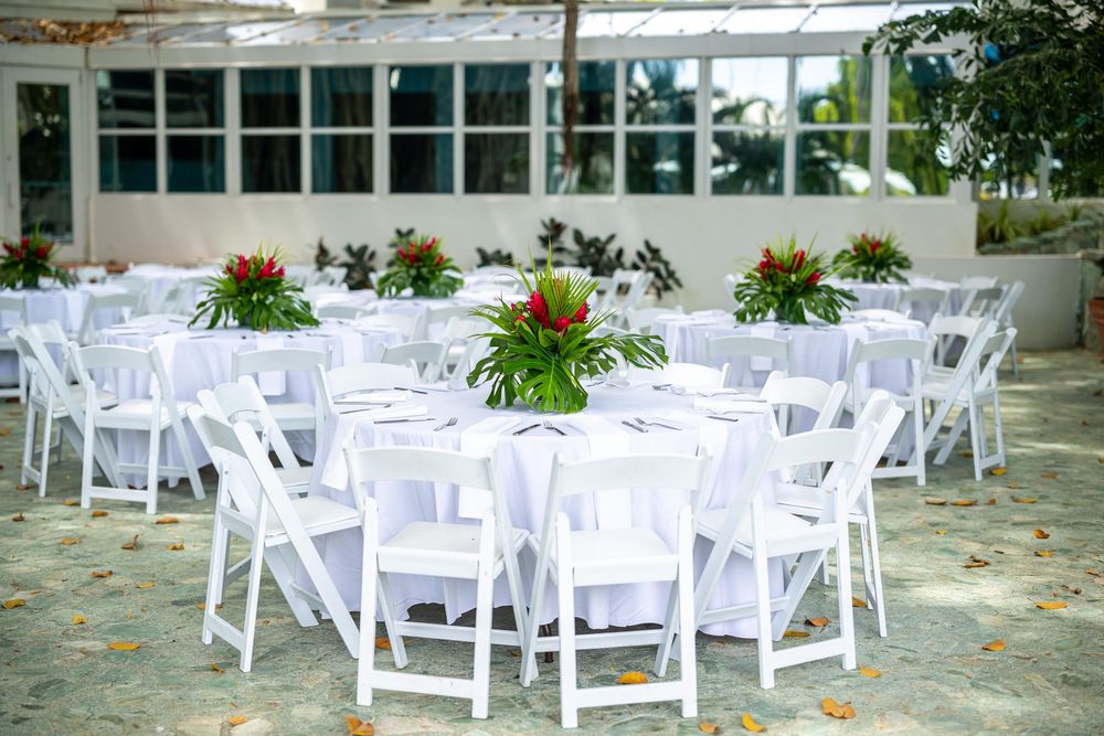 White folding chairs and round tables with tropical floral centerpieces arranged for an elegant outdoor event.