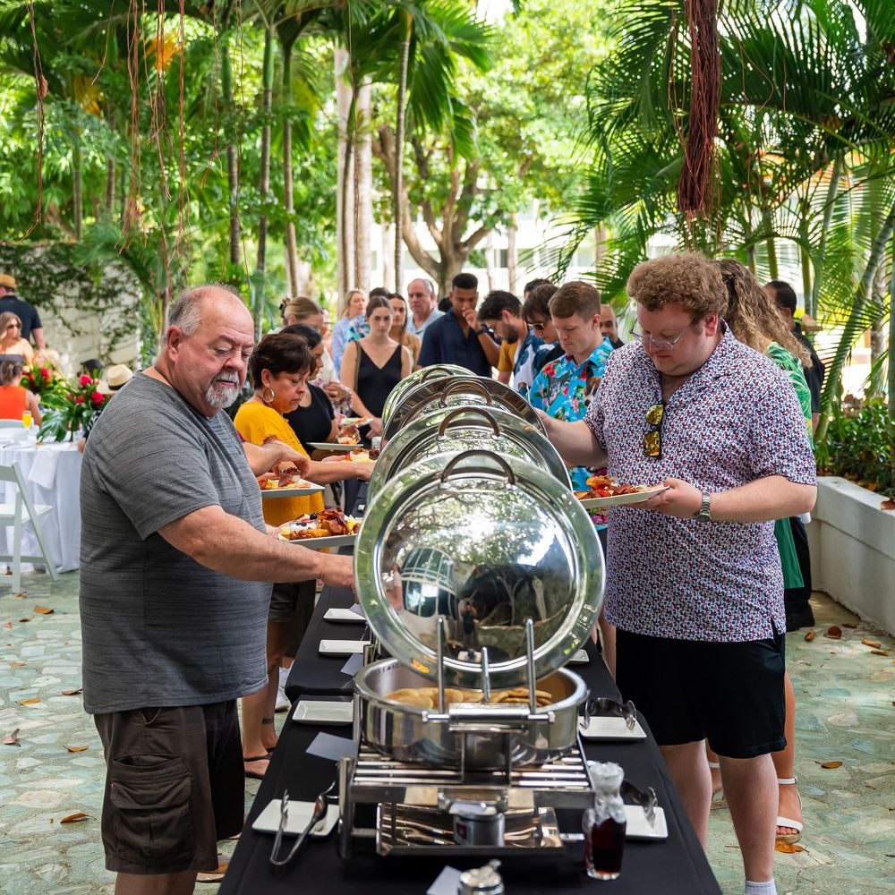 Group of people gathered around outdoor buffet tables with chafing dishes serving food in a tropical garden setting.