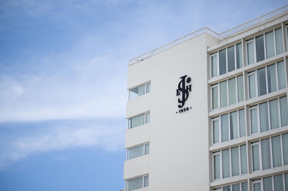A modern white hotel building with black decorative logo on its facade against a blue sky.
