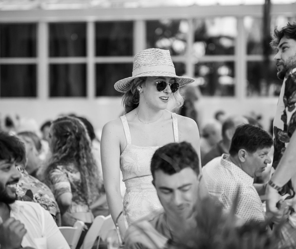 A crowd of people gathering outdoors with someone in a sun hat and sunglasses standing among them in black and white.