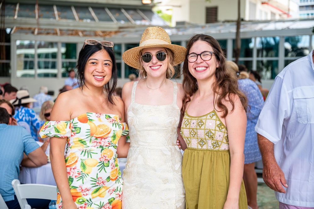 Three people in summer outfits including floral dresses and a straw hat smile together at an outdoor event.