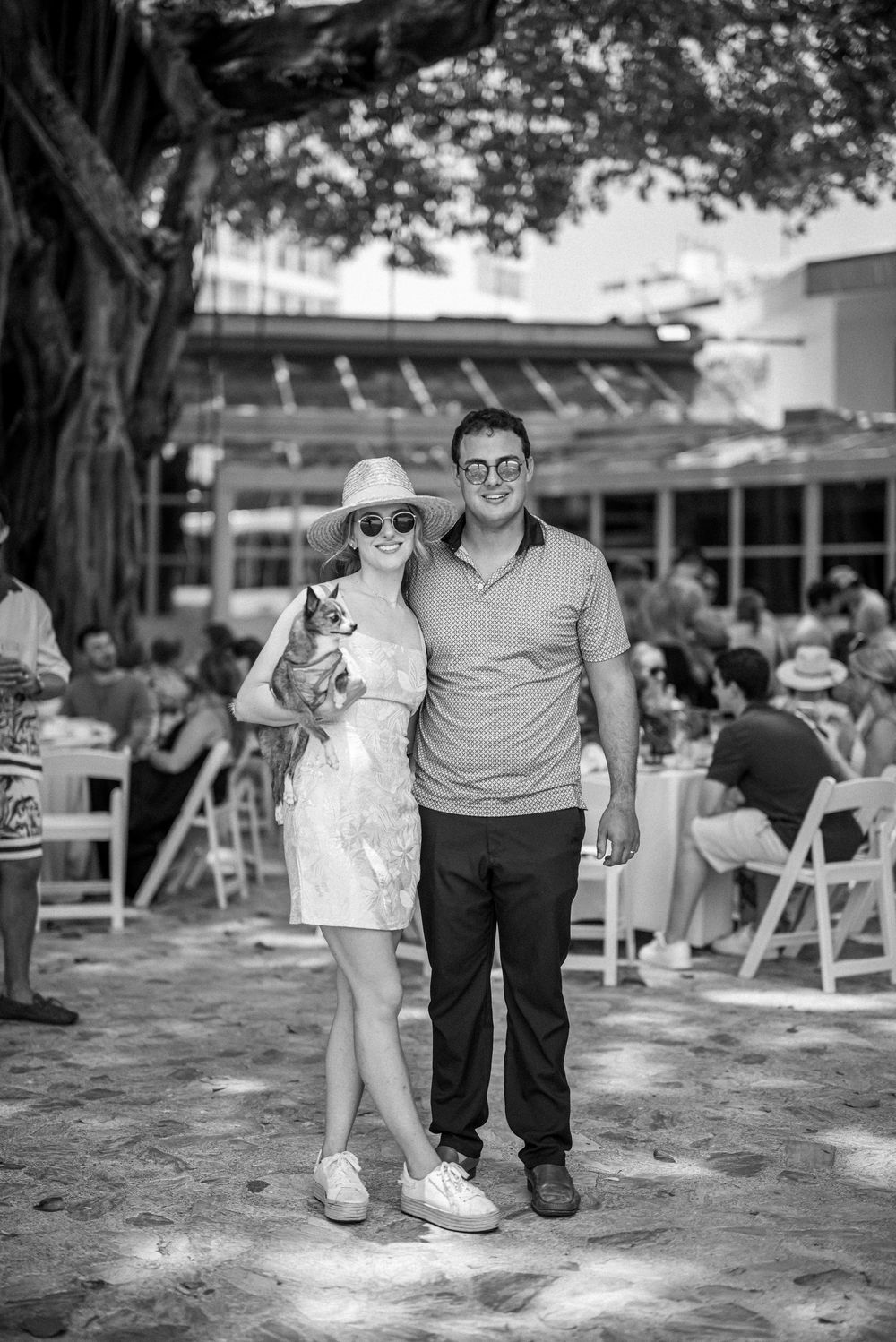 A couple stands together outdoors at a casual event venue with white chairs and tables under trees in black and white.