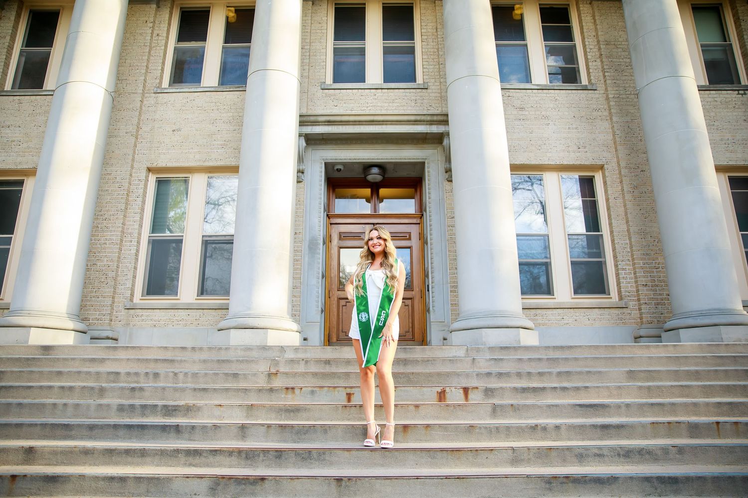 Graduate in green and white dress poses on steps of columned university building.