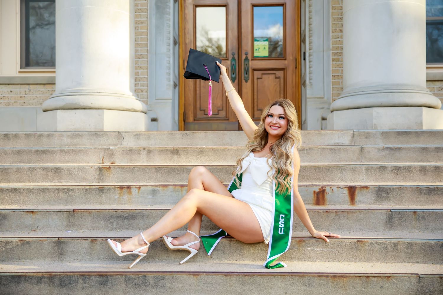 Graduate in white dress with green sash posing on stone steps of historic building.