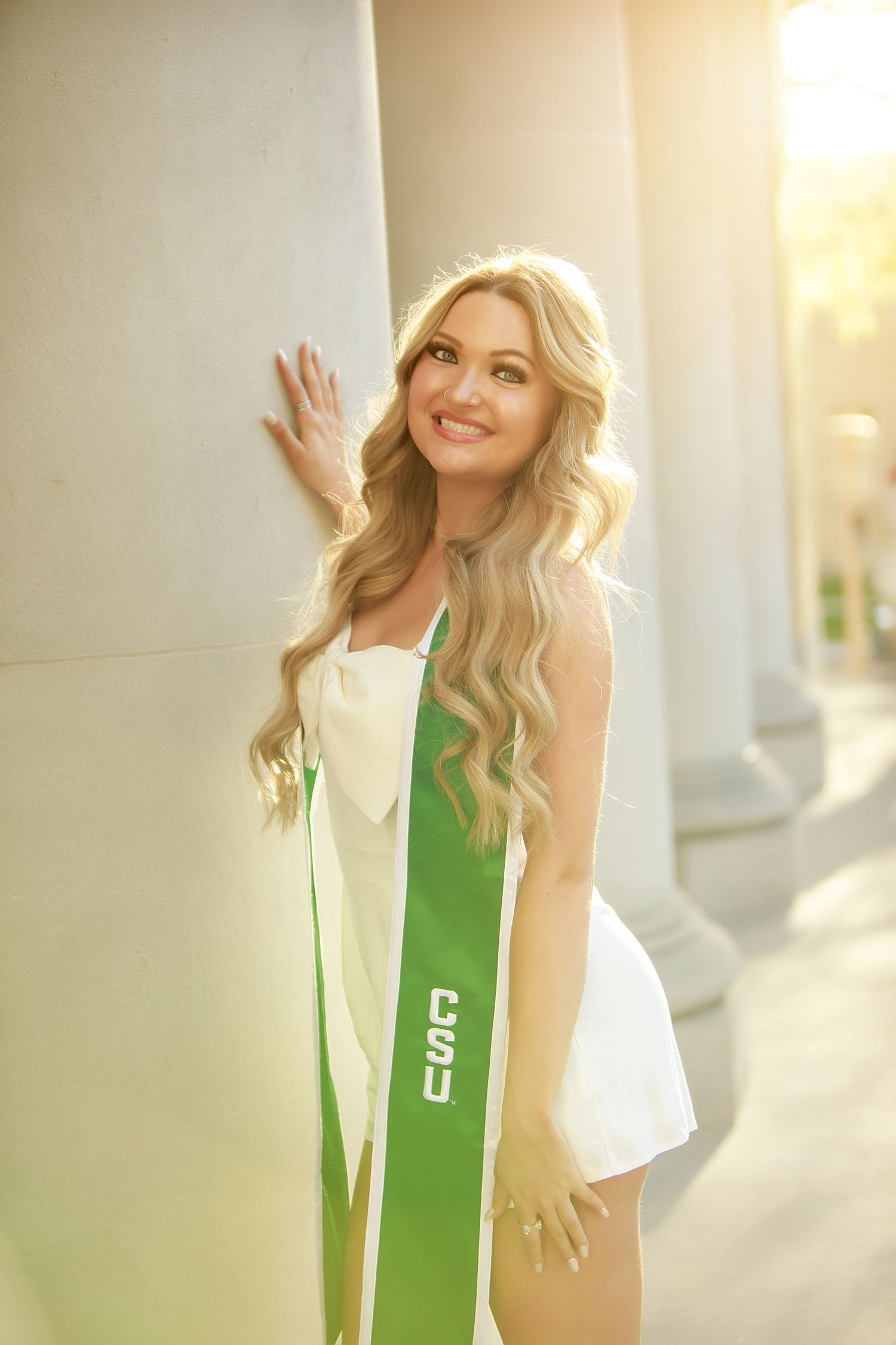 Graduate in white dress with green sash posing next to classical column in warm sunlight.