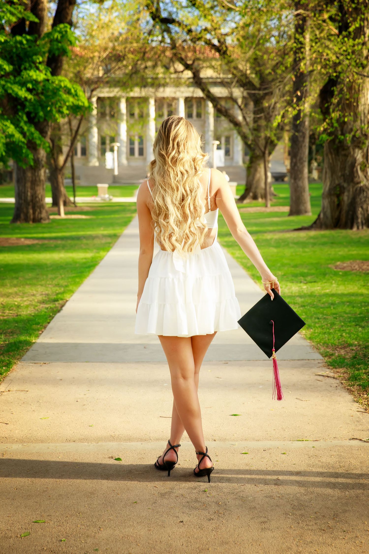 Graduate in white dress walking away on campus path holding graduation cap.
