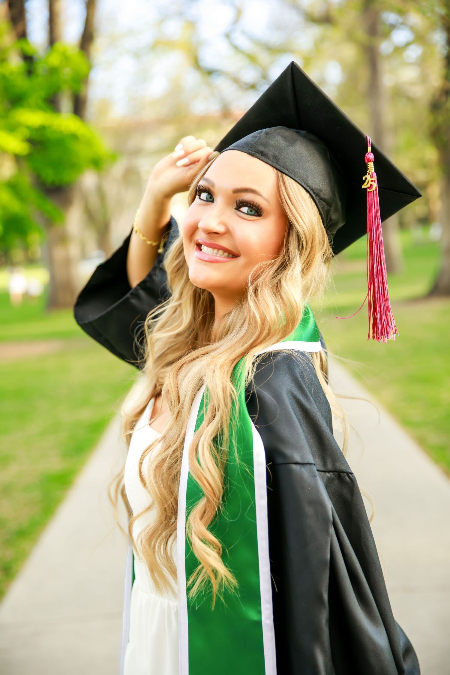 Graduate in cap and gown with green sash smiling outdoors with long blonde hair.