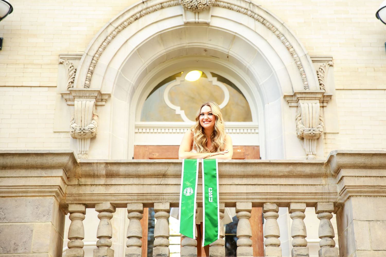 Graduate in white dress and green sash standing on classical building steps beneath ornate archway.