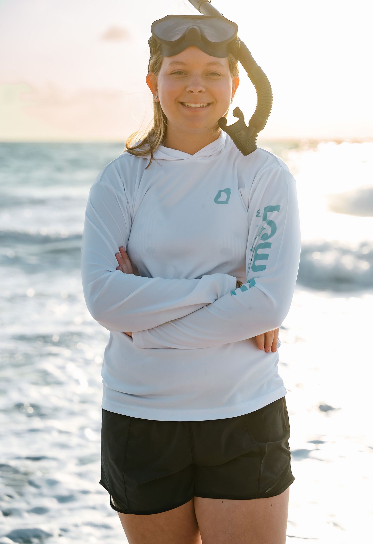 A person in a white UV protective hoodie and sunglasses stands with crossed arms at a sunny beach by the ocean.