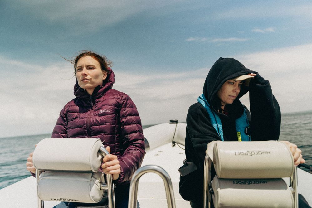 Two passengers in winter coats sit at the helm of a boat on a cloudy day at sea.
