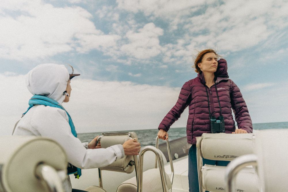 Two people having a conversation while sailing on a boat under cloudy skies with one wearing a white hood and teal scarf.