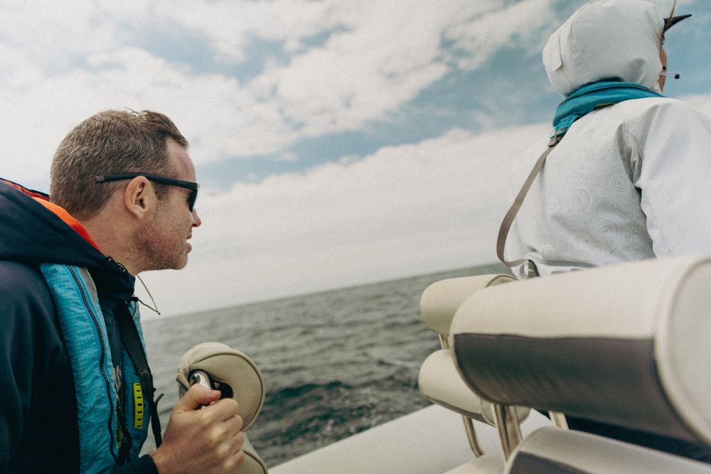 A person in a teal jacket holds a drink while sailing on a boat across open water under cloudy skies.