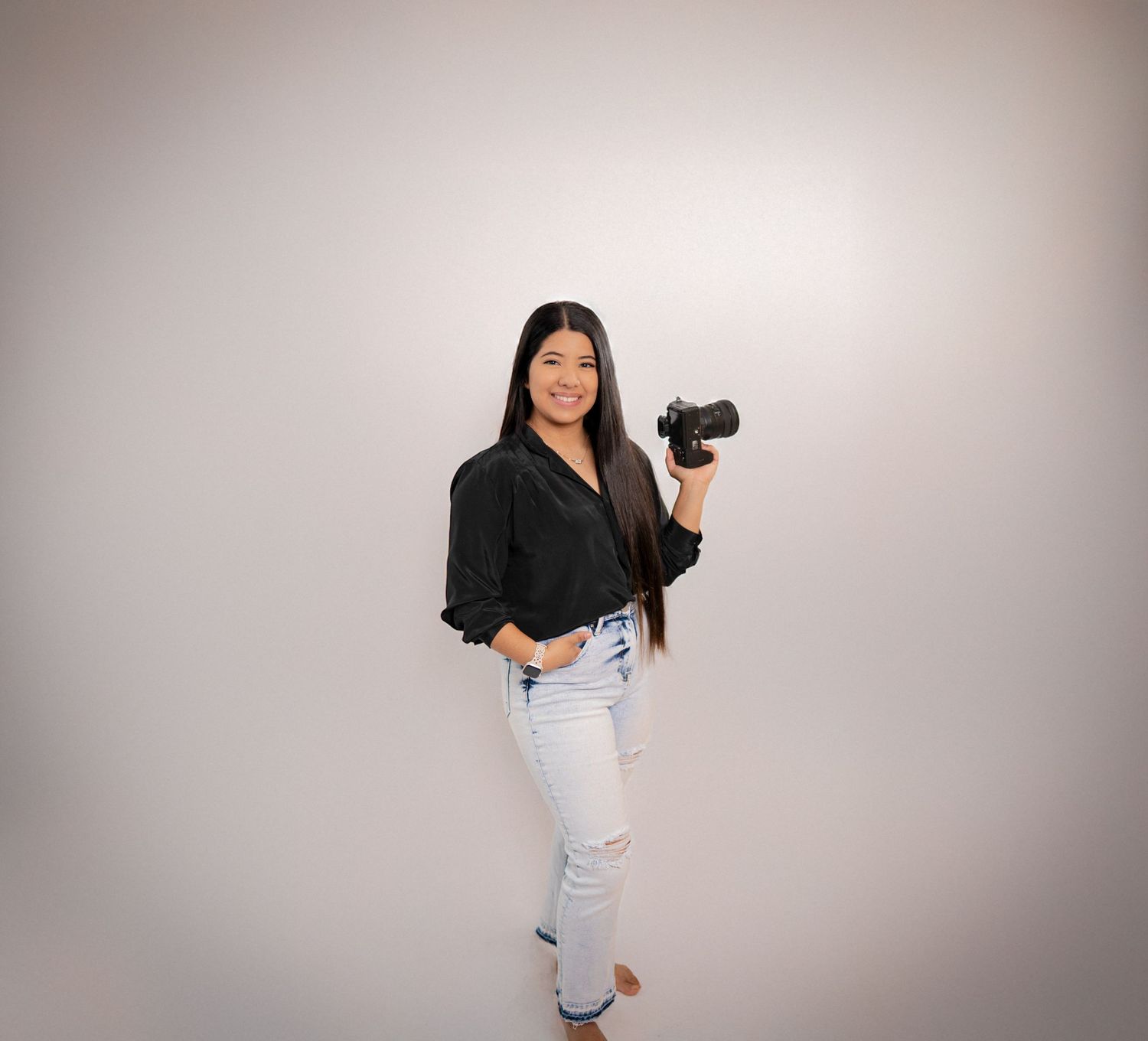 Professional photographer wearing black shirt and ripped jeans poses with a DSLR camera against studio backdrop.